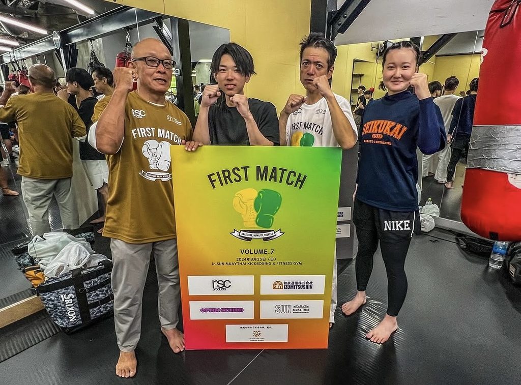 FIRST MATCH のポスターの前で仲間と記念撮影する渡邉房光さん